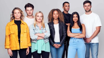 A group of seven young people of different genders and cultural backgrounds stand in a group with serious expressions wearing casual young persons' attire.