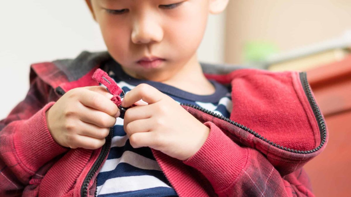 A young boy with a sombre face looks down at the zip fastener at the bottom of his jacket as he concentrates on unfastening the clasp.