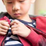 A young boy with a sombre face looks down at the zip fastener at the bottom of his jacket as he concentrates on unfastening the clasp.