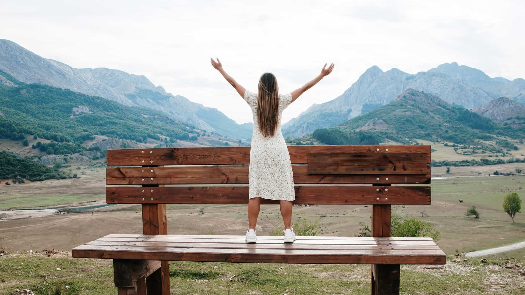 A woman stands on a huge oversized wooden park bench with her arms outstretched towards the mountainous horizon in the distance.
