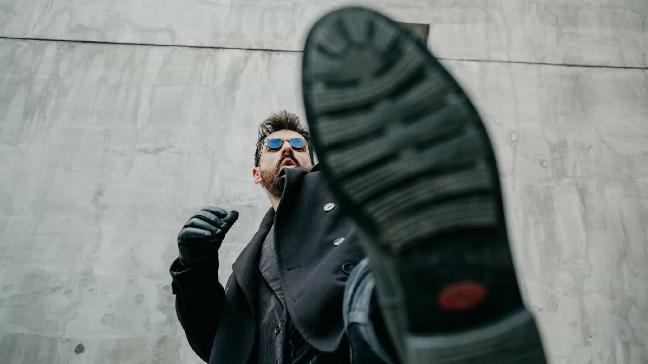 A picture taken from ground level focusing on the underside of a man's boot with the stylishly dressed man in the background wearing black amid a cold concrete background.