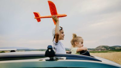 A smiling boy holds a toy plane aloft while a girl watches on from a car near an airport runway.