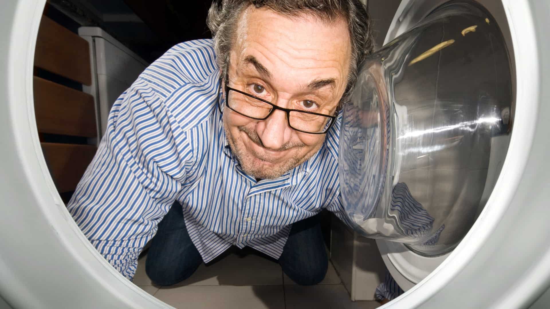 A close up of a dodgy man's face as taken from inside a washing machine as he looks in the machine with a sly grin on his face and holds the door open with one hand.