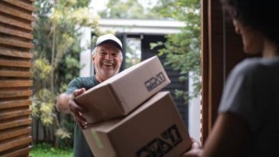 A delivery man wearing a cap and smiling broadly delivers two boxes stacked on top of each other at the door of a female customer whose back can be seen at the edge of a doorway.