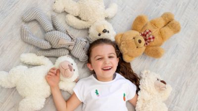 A cute young girl lays on the floor with five teddy bears lying in a semicircle head to head with her as she clutches another teddy bear in one arm.