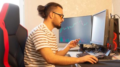 A hip young guy works at his home workstation with two screens and a gamers chair, keeping an eye on his crypto investments.