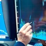 A man sits with his head in his hand, looking quite dejected, as he holds a rubber tipped pen on the screen of a computer showing a graph trending downwards.