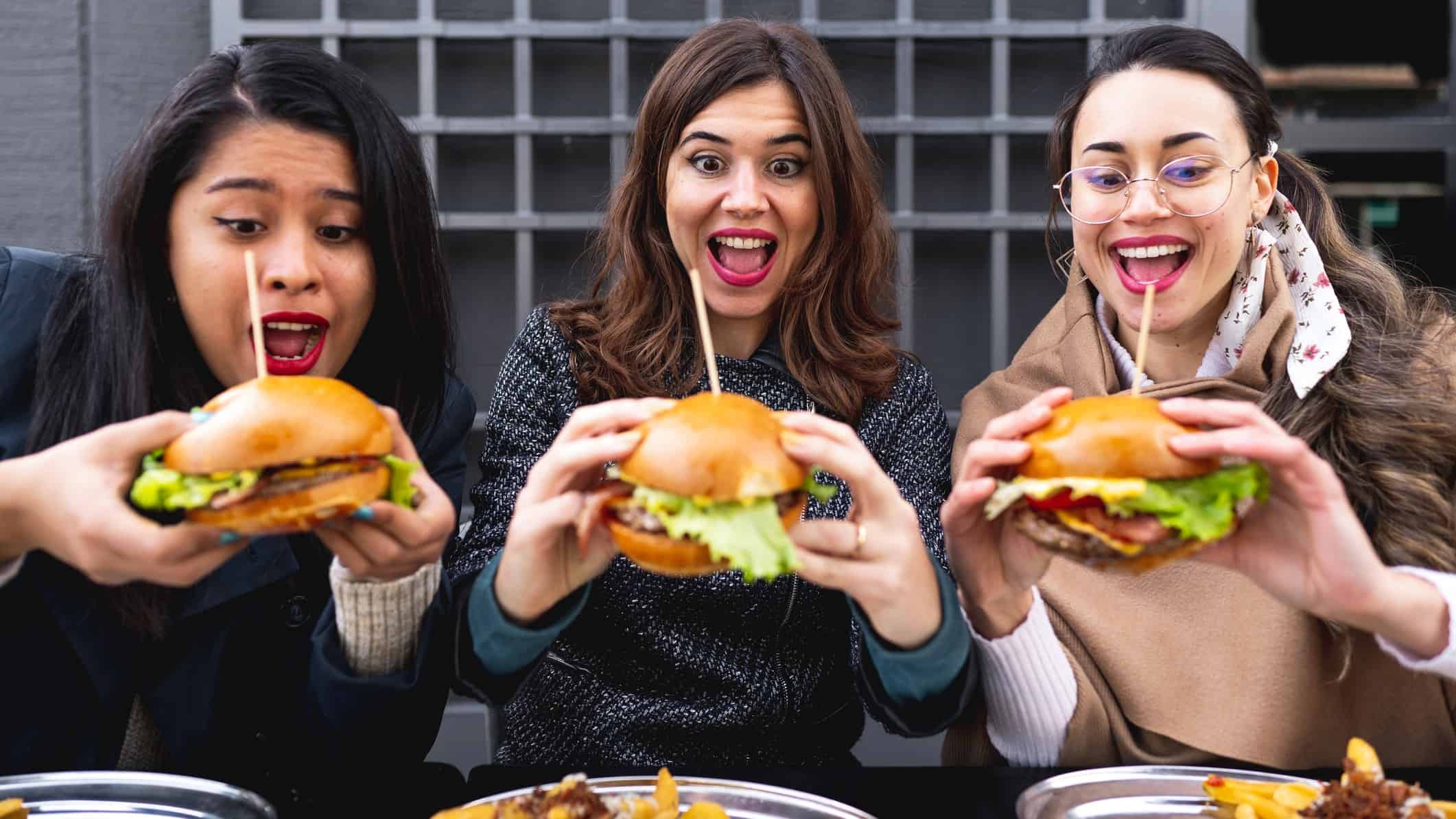 Three young women sit side by side each holding large hamburgers with the lot skewered with bamboo sticks in their two hands with wide, happy smiles on their faces as they look at their hamburgers just before they are about to tuck into them.