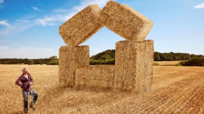 a young farmer stands back and admires his work in arranging bales of hay to form a house shape with two bales balancing against each other to form a roof, perched on bales tipped on their side in an abstract house shape on a freshly harvested paddock.