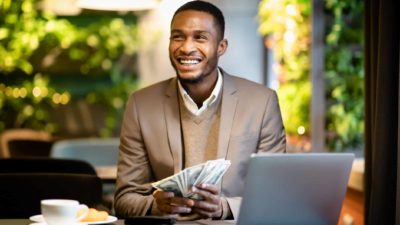 A man smiles as he holds bank notes in front of a laptop.