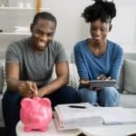 A couple sits in their lounge room with a large piggy bank on the coffee table. They smile while the male partner feeds some money into the slot while the female partner looks on with an iPad style device in her hands as though they are budgeting.