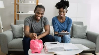 A couple sits in their lounge room with a large piggy bank on the coffee table. They smile while the male partner feeds some money into the slot while the female partner looks on with an iPad style device in her hands as though they are budgeting.