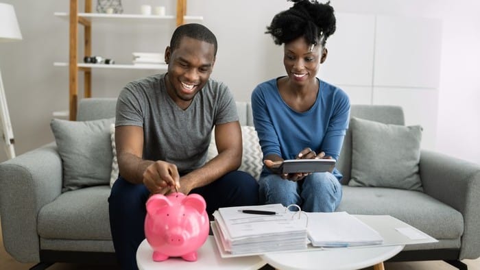 A couple sits in their lounge room with a large piggy bank on the coffee table. They smile while the male partner feeds some money into the slot while the female partner looks on with an iPad style device in her hands as though they are budgeting.