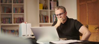 A senior investor wearing glasses sits at his desk and works on his ASX shares portfolio on his laptop.