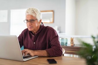 A senior investor wearing glasses sits at his desk and works on his ASX shares portfolio on his laptop2