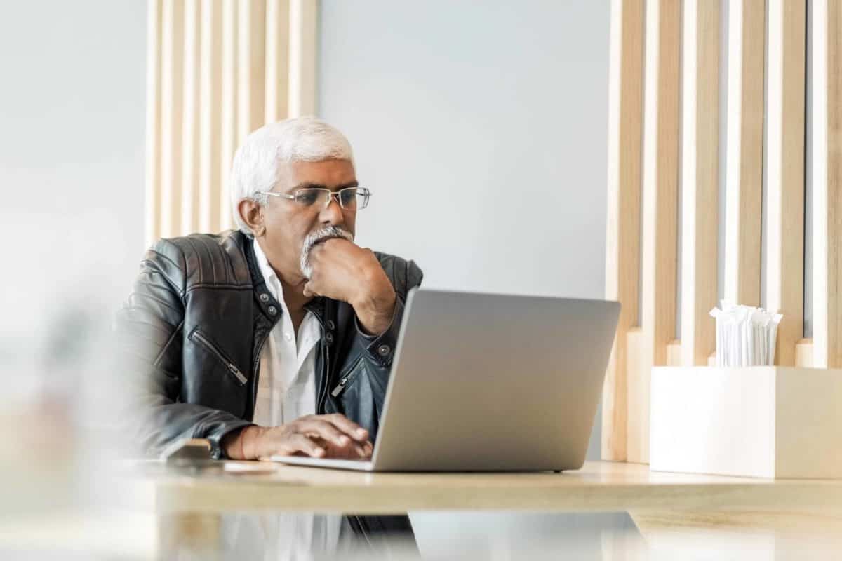 Senior man wearing glasses and a leather jacket works on his laptop in a cafe.