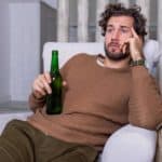A young man holds a small bottle of beer as he slumps sadly on one elbow in a comfortable chair with his head propped in his hand and staring into space with a dejected look on his face.