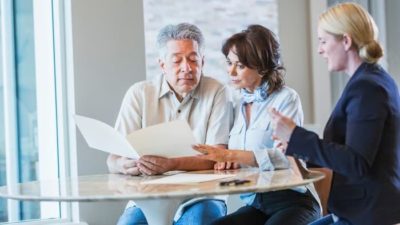 A senior couple sets at a table looking at documents as a professional looking woman sits alongside them as if giving retirement and investing advice.