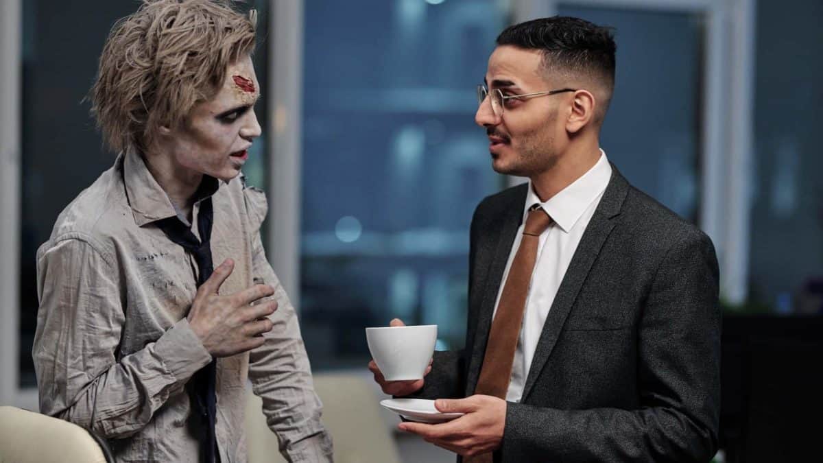 A businessman holding a cupof tea chats to a zombie in the office.