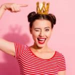 A woman weraing a stripy t-shirt winks as she points to the decorative gold crown on her head.