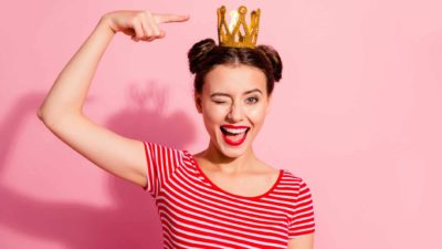 A woman weraing a stripy t-shirt winks as she points to the decorative gold crown on her head .