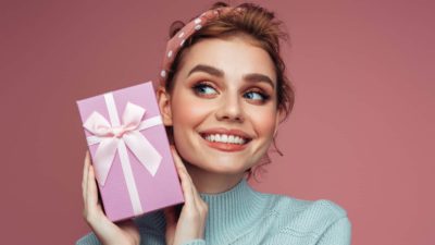 A girl smiles broadly as she holds a gift box complete with ribbon up to her face as though shaking it to guess what's inside.