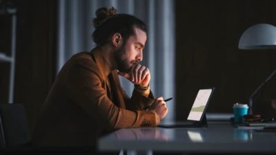 A hip young man with a beard and manbun sits thoughtfully at his laptop computer in a darkened room, staring at the screen with his chin resting on his hand in thought.
