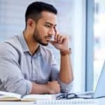 A man in his 30s with a clipped beard sits at his laptop on a desk with one finger to the side of his face and his chin resting on his thumb as he looks concerned while staring at his computer screen.