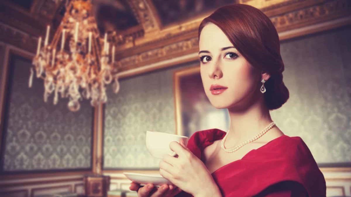 A formally dressed young woman sips tea from a china cup and saucer as she gives a haughty look against the background of a European style drawing room with heavy wood, traditional wallpaper and a large chandelier hanging from the ceiling.