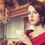 A formally dressed young woman sips tea from a china cup and saucer as she gives a haughty look against the background of a European style drawing room with heavy wood, traditional wallpaper and a large chandelier hanging from the ceiling.