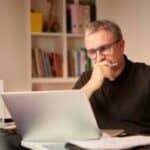 A senior investor wearing glasses sits at his desk and works on his ASX shares portfolio on his laptop.