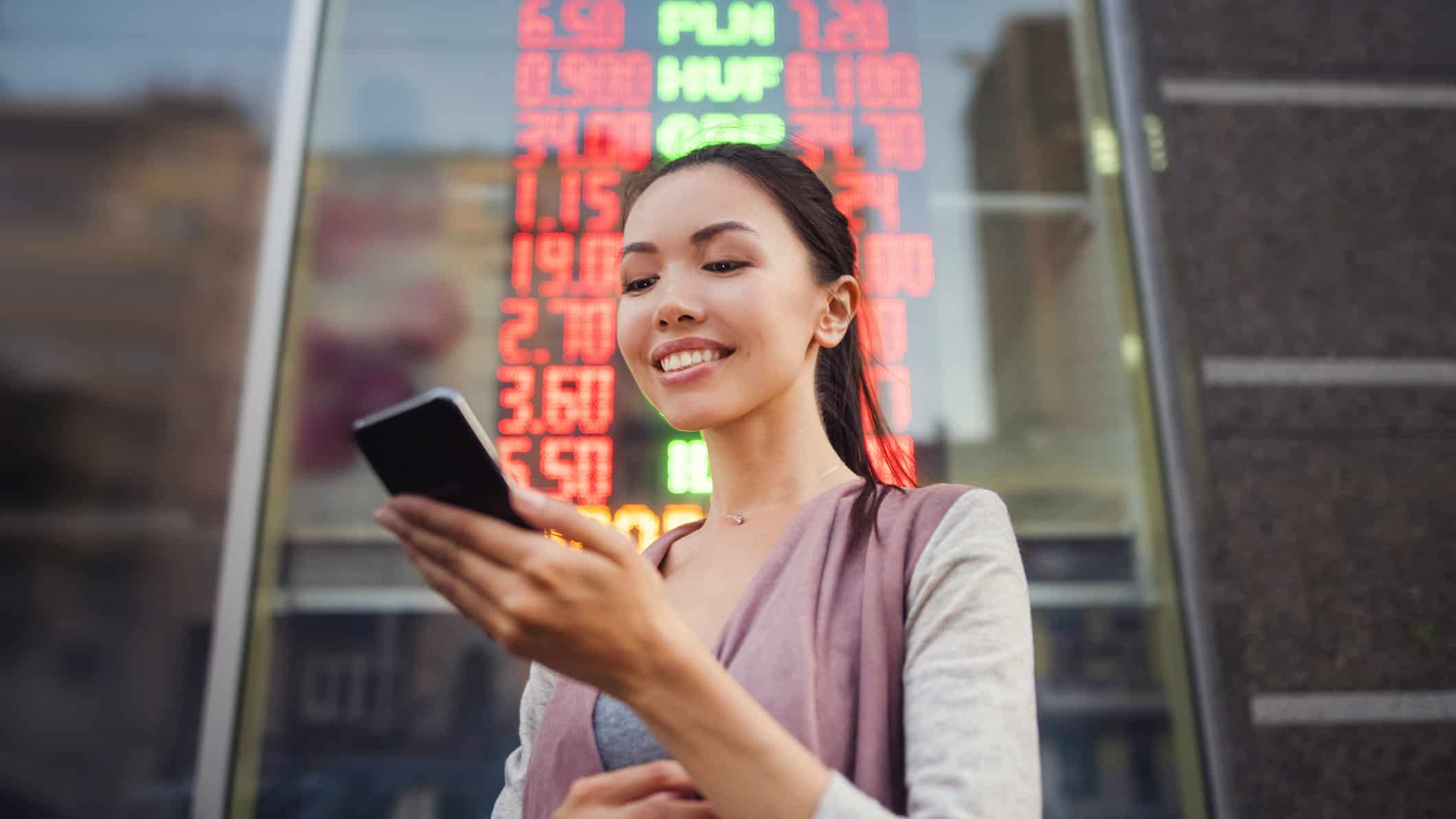 A young woman uses an application in her smart phone to check currency exchange rates in front of an illuminated information board.