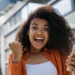 A young woman holding her phone smiles broadly and looks excited, after receiving good news.