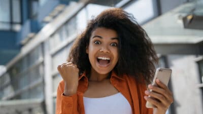 A young woman holding her phone smiles broadly and looks excited, after receiving good news.