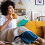 A woman relaxes on a yellow couch with a book and cuppa, and looks pensively away as she contemplates the joy of earning passive income.