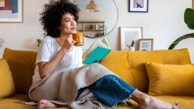 A woman relaxes on a yellow couch with a book and cuppa, and looks pensively away as she contemplates the joy of earning passive income.