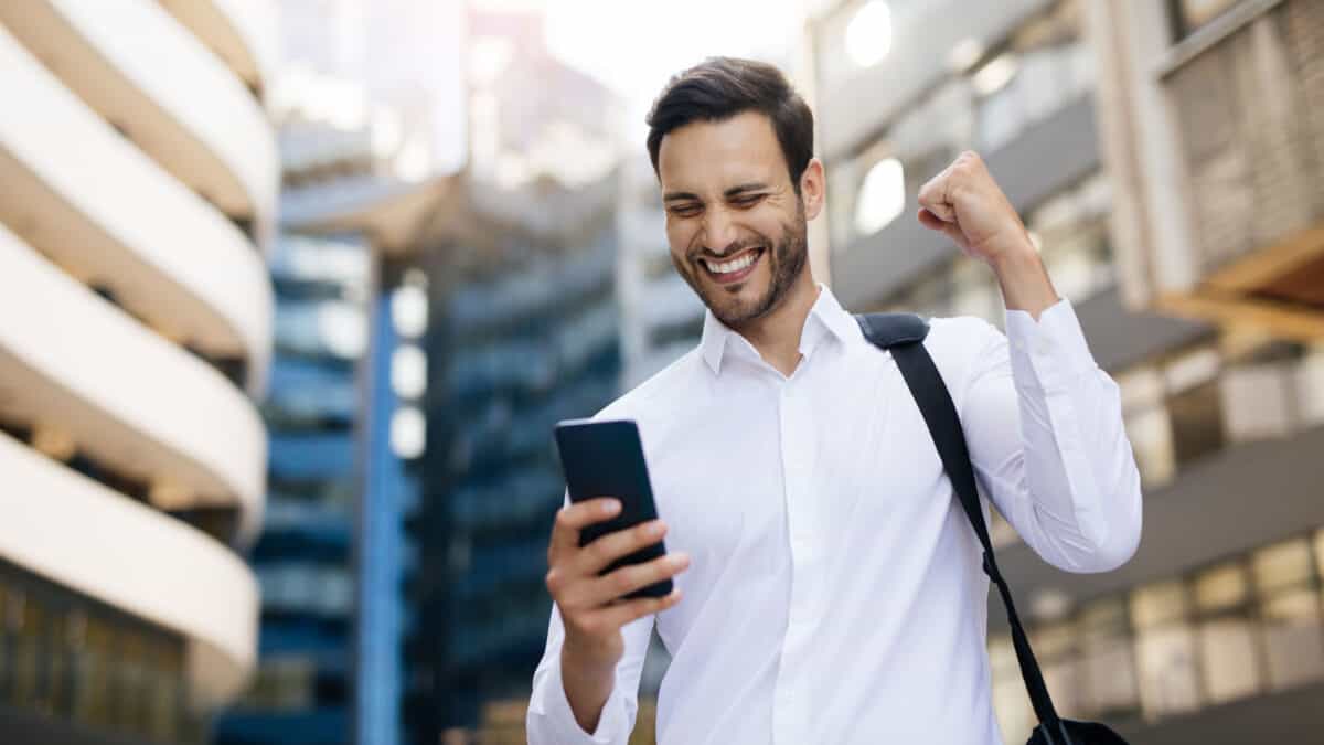 A smiling businessman in the city looks at his phone and punches the air in celebration of good news.