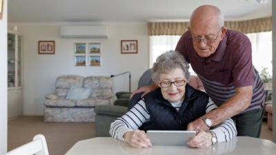An older gentleman leans over his partner's shoulder as she looks at a tablet device while seated at a table in their classic Australian old person's home, complete with comfortable furniture and family photographs on the walls.
