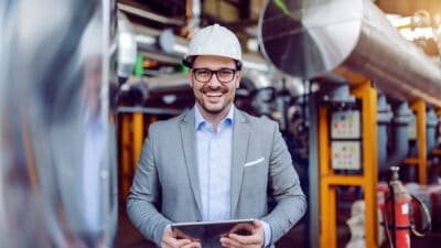 Smiling attractive caucasian supervisor in grey suit and with white helmet on head holding tablet while standing in power plant.