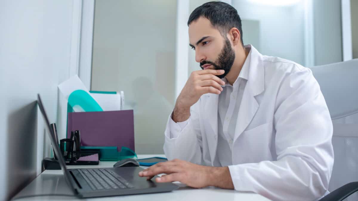 Male doctor in a lab coat working at laptop looking serious.