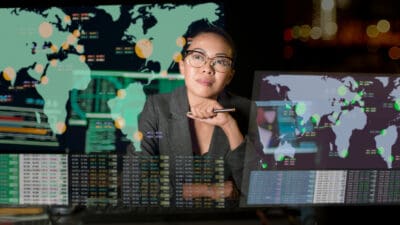 A woman sits at her desk thinking. She is surrounded by projections of world maps on various screens with data appearing below them.