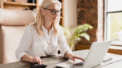 Woman calculating dividends on calculator and working on a laptop.