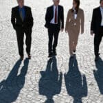 Four business people wearing formal business suits and ties walk abreast on a wide paved surface with their long shadows falling on the ground ahead of them.