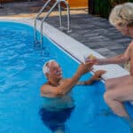 Loving senior couple at the swimming pool. Senior woman sitting on the edge of the pool and giving lemonade to her husband who is swimming.