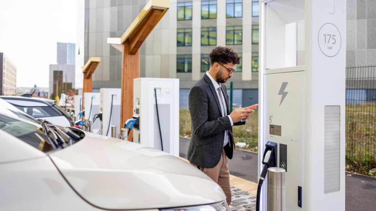 A man checks his phone next to an electric vehicle charging station with his electric vehicle parked in the charging bay.