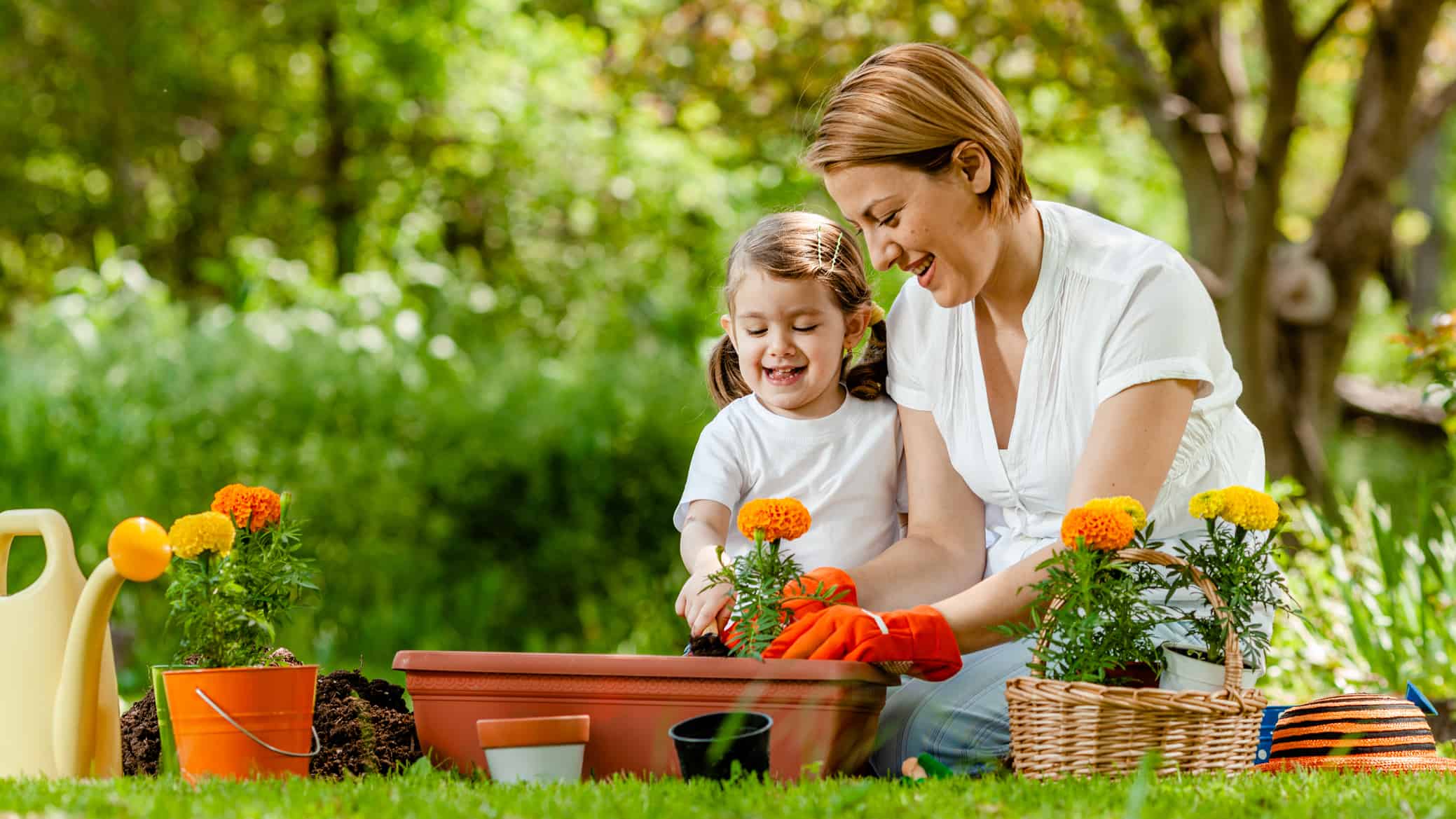 A woman and her child plant flower seedlings in a planter box in a green garden setting.