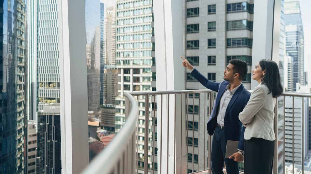 A man and a woman stand on an external balcony in a dense city environment filled with high rise buildings and commercial properties. The man is pointing up at a high rise building and the woman is looking on.