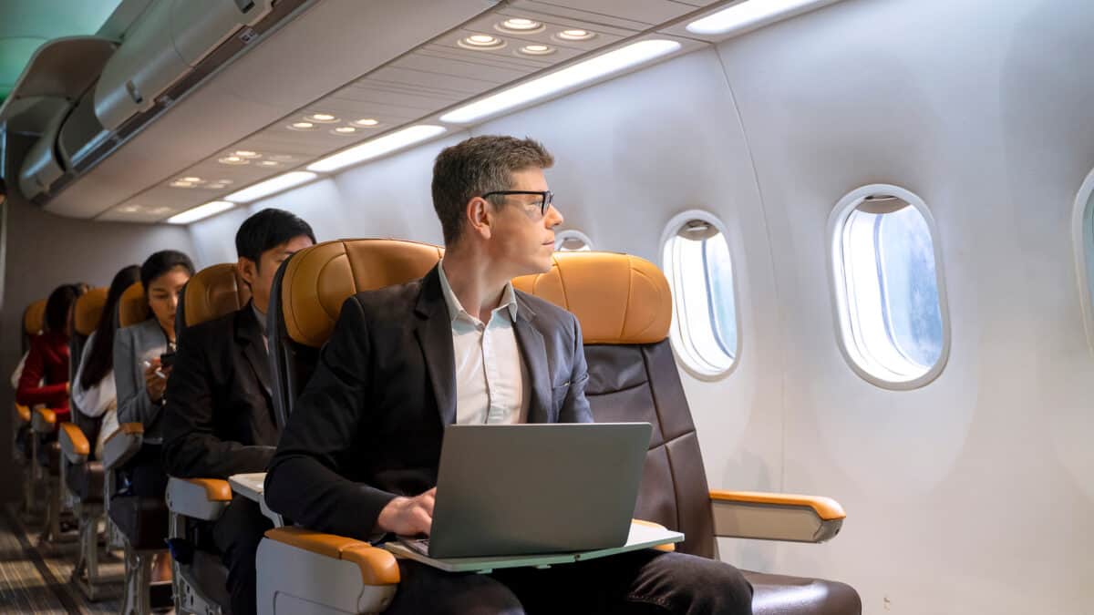 Man sitting in a plane looking through a window and working on a laptop.