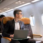 Man sitting in a plane looking through a window and working on a laptop.