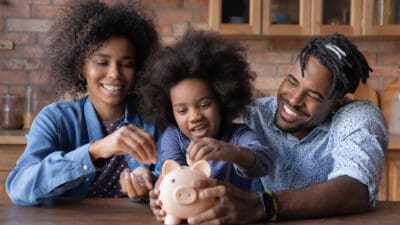 Young girl starting investing by putting a coin ion a piggybank while surrounded by her parents.
